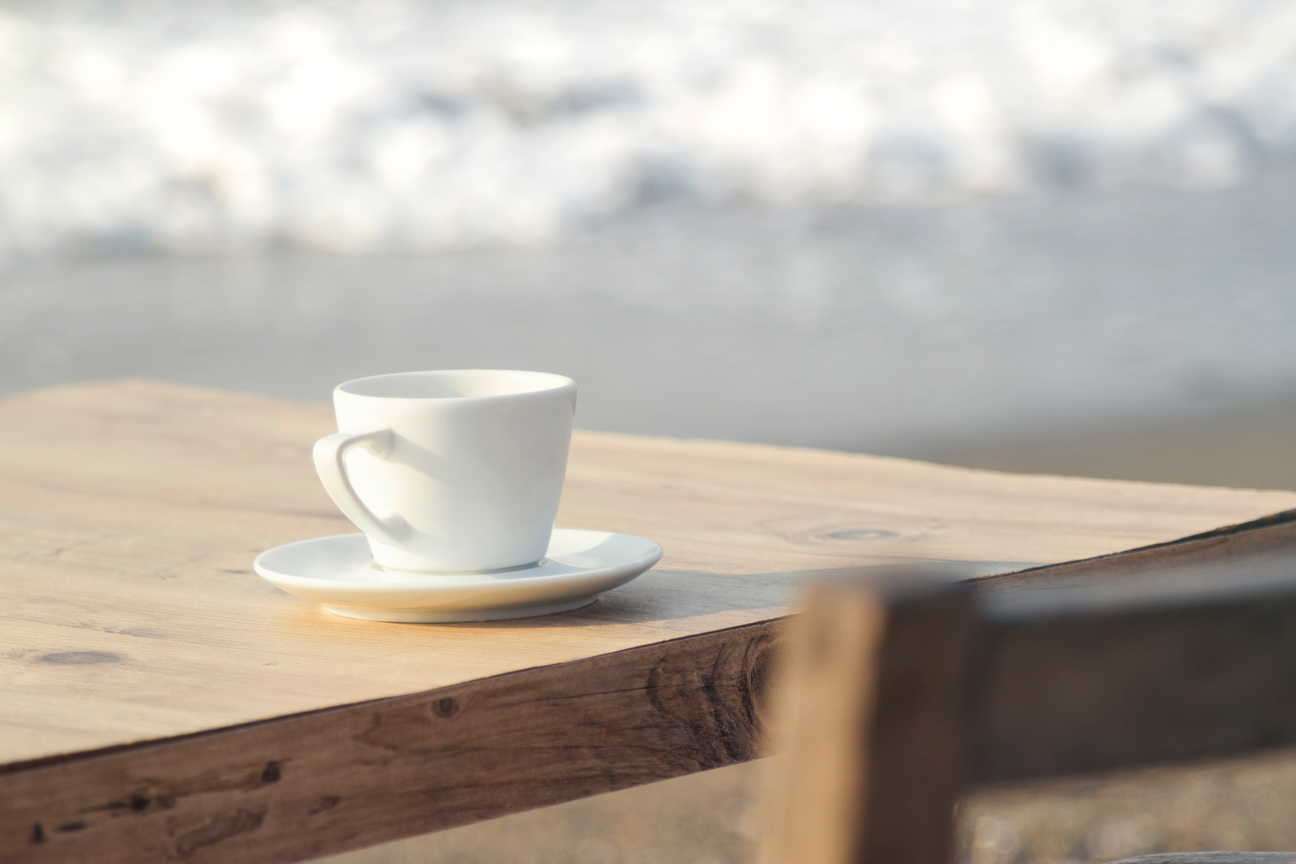 A cup of coffee with a view: a steaming mug sits on a wooden table beside the ocean.